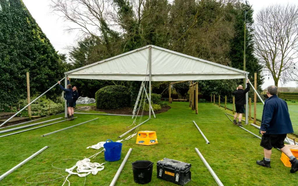 Three people assembling a large white marquee tent in a grassy area.