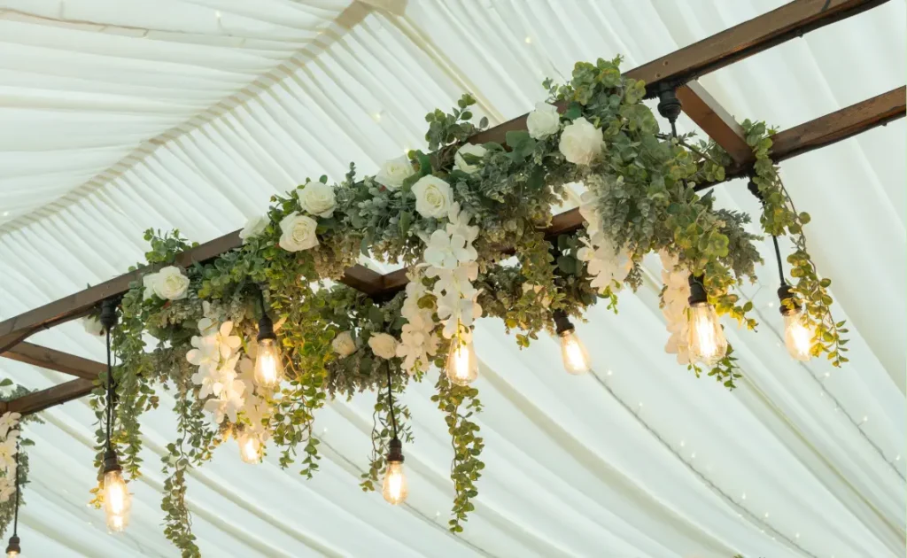 Elegant floral arrangement with white roses and greenery draped over a wooden beam, adorned with hanging lights in a marquee with white drapery.