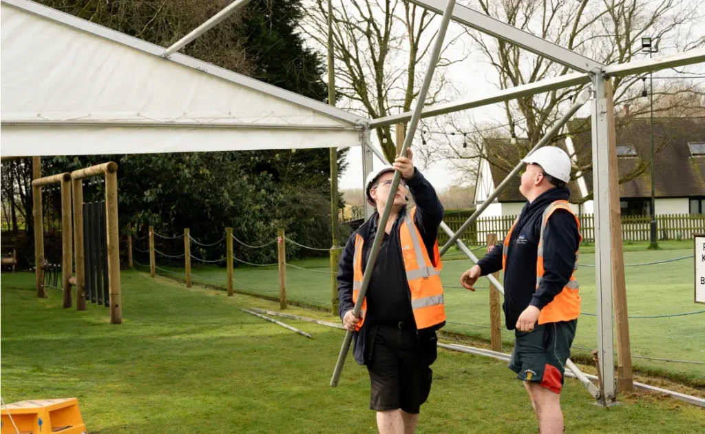 Two workers in high-visibility vests assemble a metal frame for a marquee outdoors.