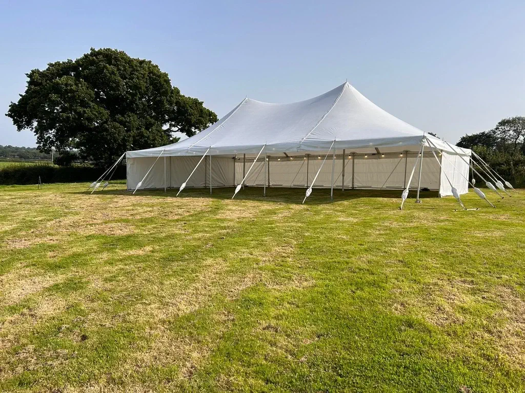 A large white traditional marquee is set up on a grassy field, secured with multiple ropes. Trees are visible in the background under a clear sky.