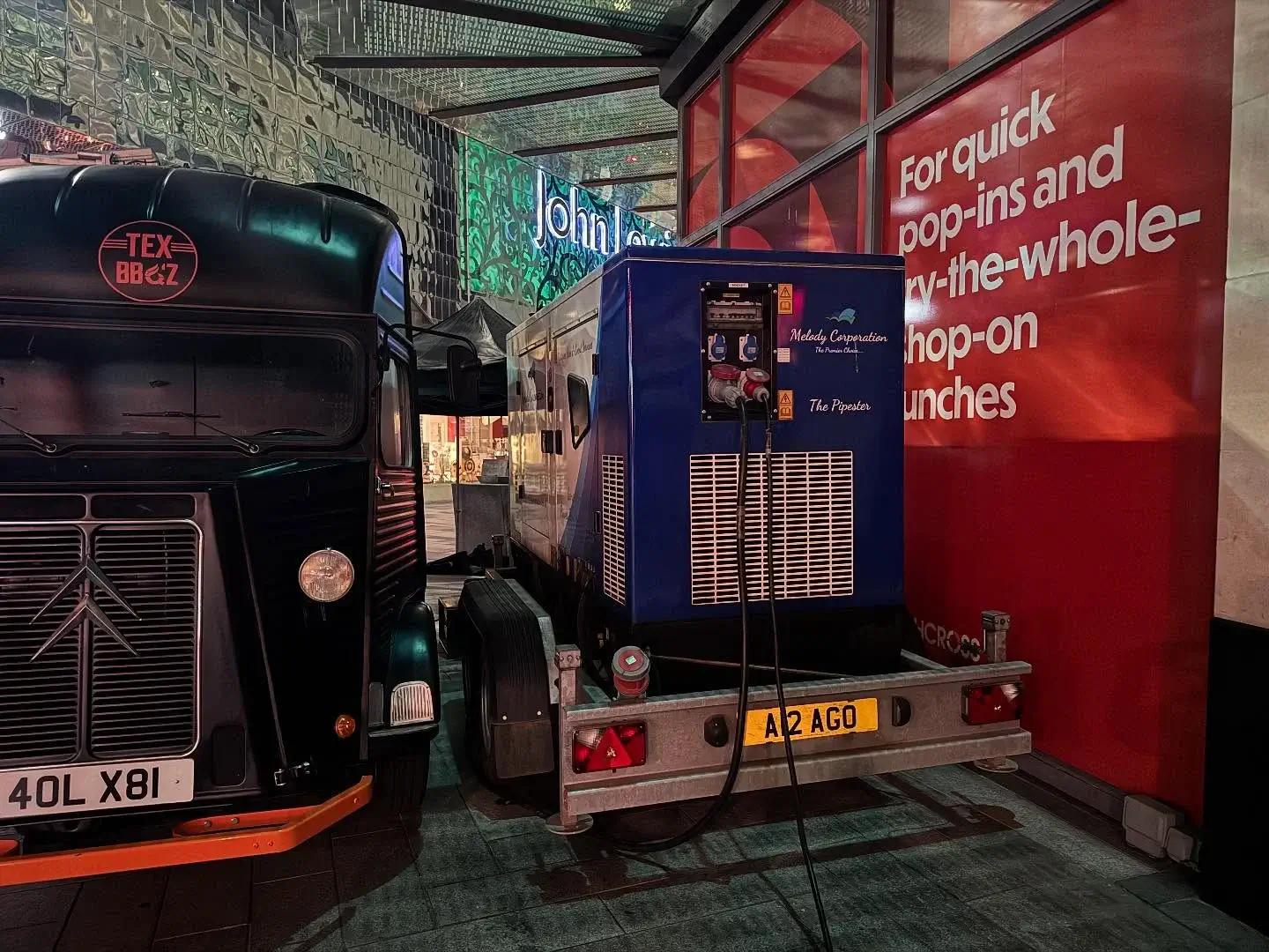 A blue mobile generator on a trailer is parked next to a black van near a red wall with white text promoting quick lunch options.