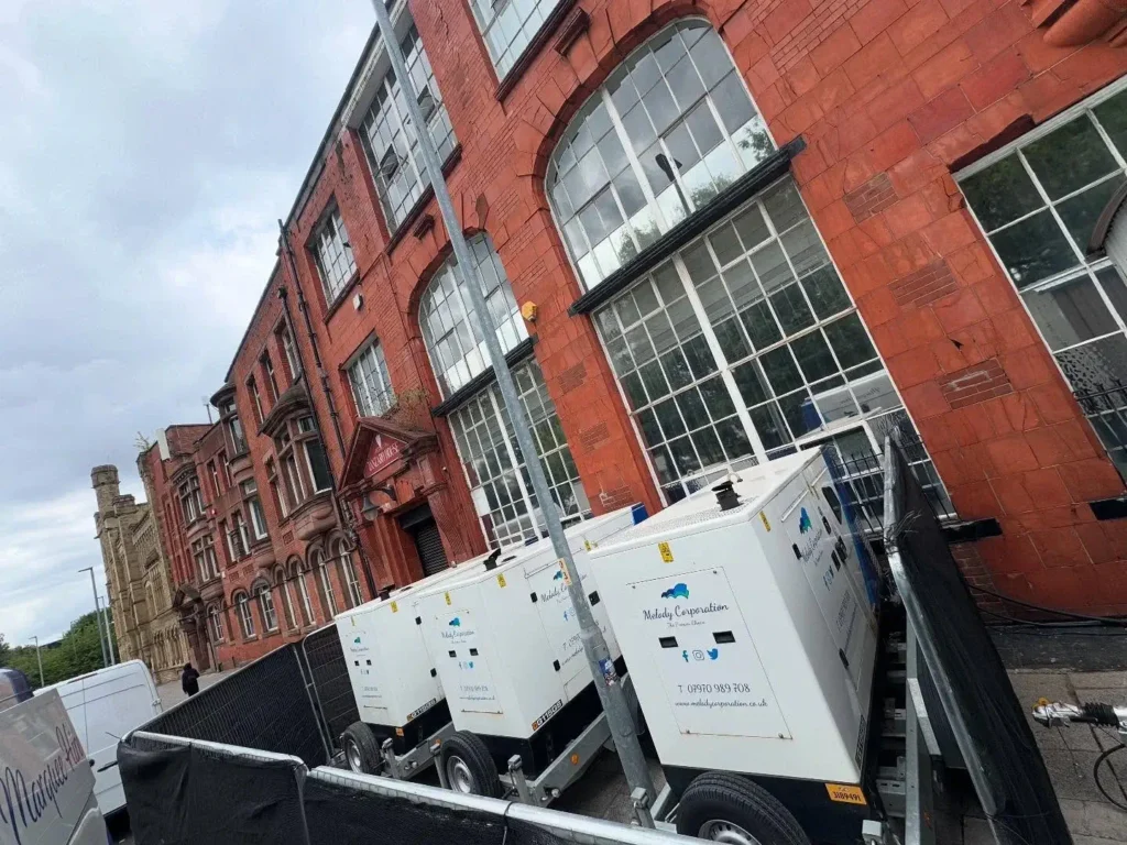 Two large industrial generator units are placed behind a metal fence outside a red-brick building with large windows on a cloudy day.