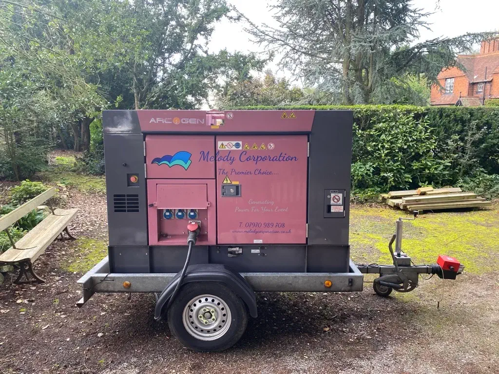 A portable generator on a trailer sits outdoors, surrounded by trees and benches, with contact information displayed on its side.