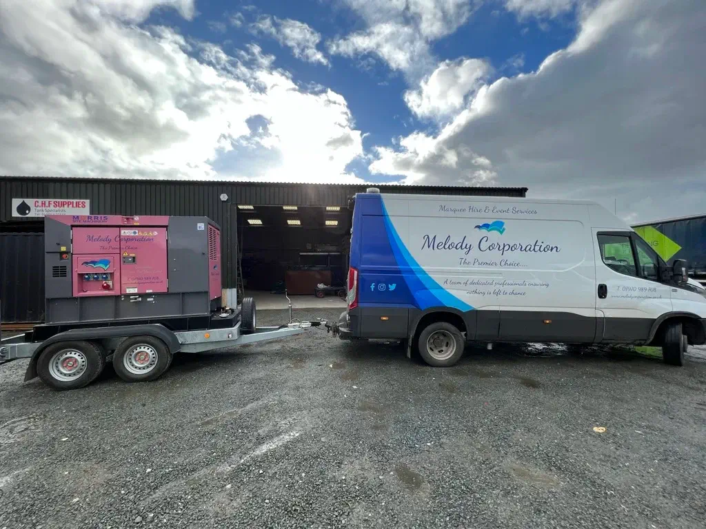 A white Melody Corporation van is parked outside a warehouse, towing a trailer with a large pink generator on a gravel surface under a cloudy sky.