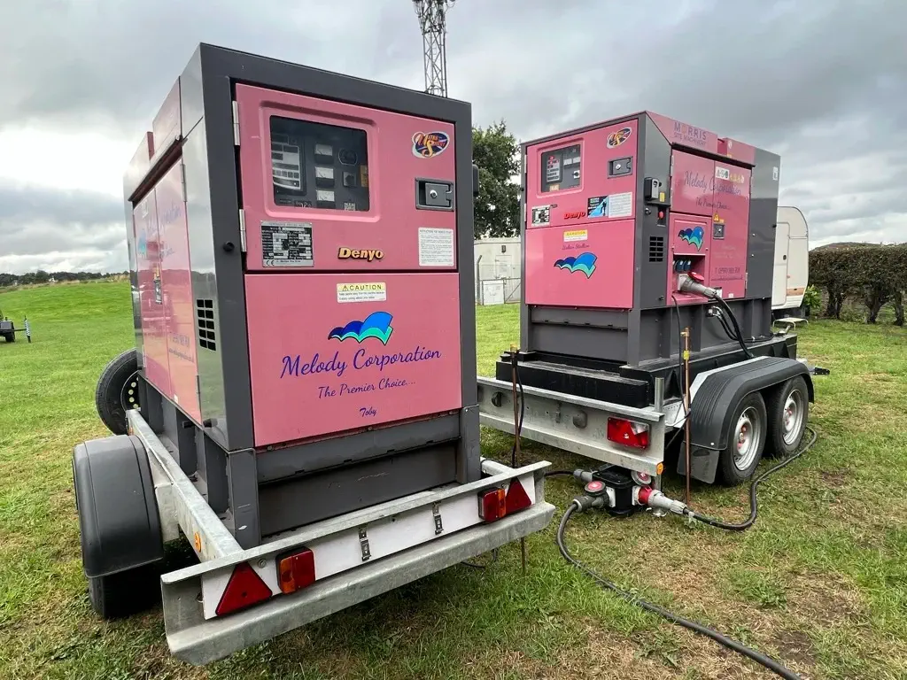 Two pink industrial generators labeled "Melody Corporation" are mounted on trailers and set up on grass, connected by cables.
